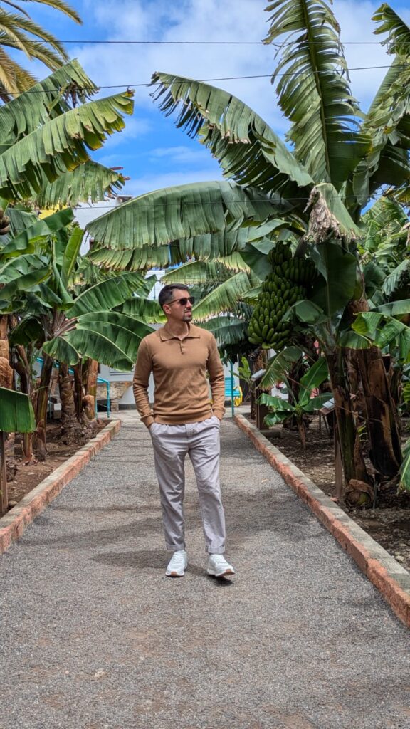 Un hombre joven con gafas de sol y polo color ocre camina por un sendero flanqueado por plataneras bajo un cielo azul despejado.