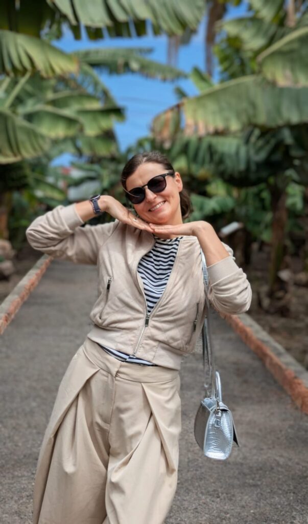 Una mujer sonriente con gafas de sol posa en un sendero rodeado de plataneros, apoyando la barbilla sobre sus manos en un gesto alegre.